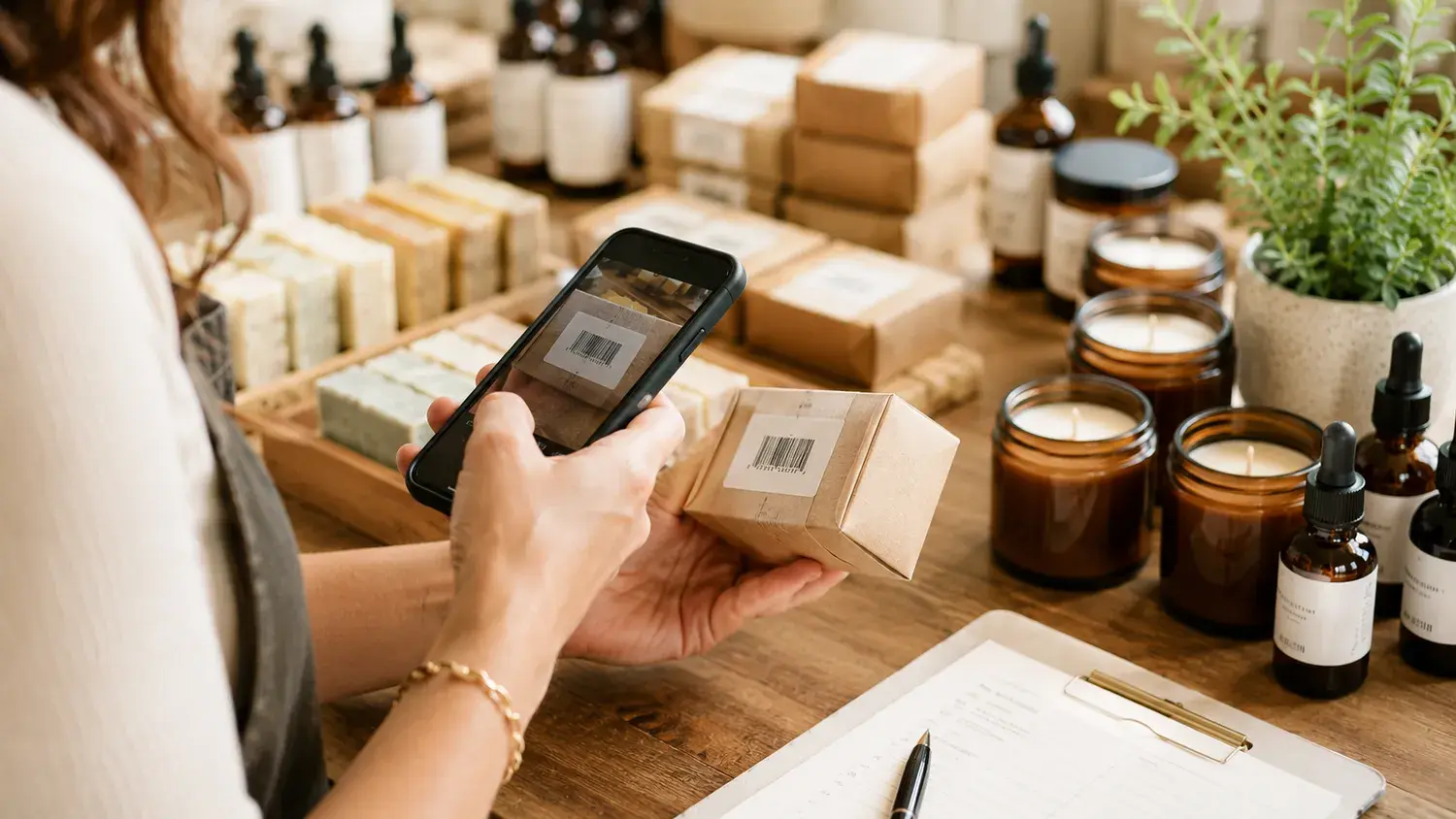 A maker scanning the barcode on a kraft shipping box with her phone camera, candles and soaps on the table